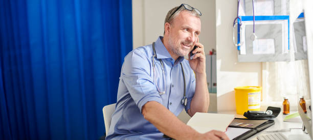 Doctor phone consultation mature GP at his desk rolled up sleeves stock pictures, royalty-free photos & images