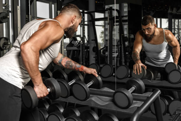 istock Bodybuilding. Bearded man exercising at gym with dumbbell looking at mirror checking technique side view 1158915033