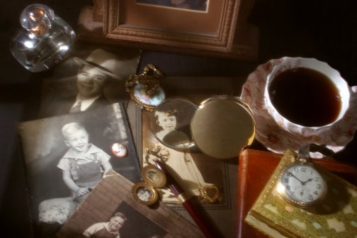 Collection of vintage photographs, one highlighted through a magnifying glass.  Cup of coffee, old pocket watch, locket, necklace, choaker locket, perfume bottle,  cameo and fountain pen.  Diffused, soft lighting creates the scene and atmosphere. Retro, old-fashioned. 