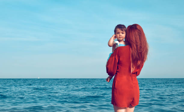 moments in vacation mother with her baby girl embracing and looking at sea in summer day. little redhead girl stock pictures, royalty-free photos & images