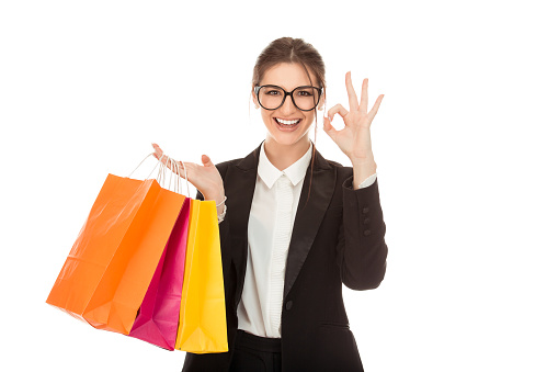 Happy young woman wearing formal white buttoned shirt and black suit jacket standing isolated over pure white background, carrying shopping bags, showing ok sign gesture with hand