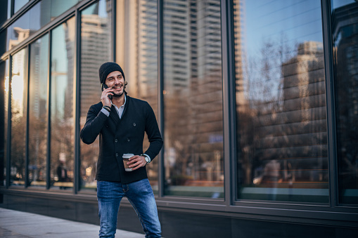 Handsome man in casual clothing standing on the street, holding takeout coffee and talking on smart phone.