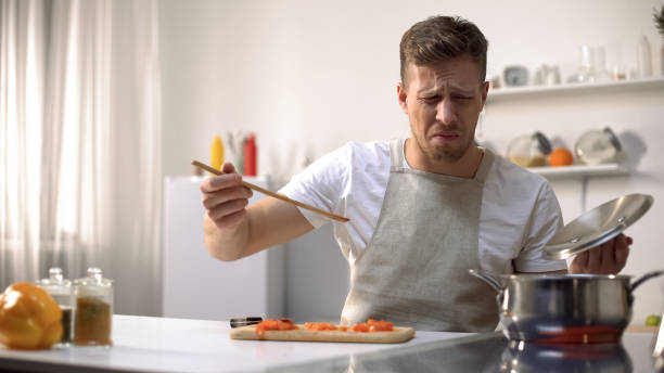 young man tasting cooked food with disgusted face expression, funny grimacing - offense imagens e fotografias de stock