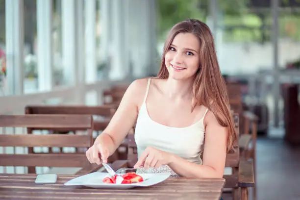 Photo of Young woman wearing white shirt, casual clothes looking at you camera smiling while eating her healthy salad food sitting on a terrace of a  trendy cafe