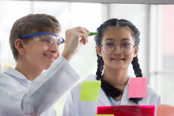 Photo of Pupils in white gown doing a chemical experiment write on glass board in laboratory at school  . kids in science lab study . multi ethnic . asian , caucasian , biochemistry .