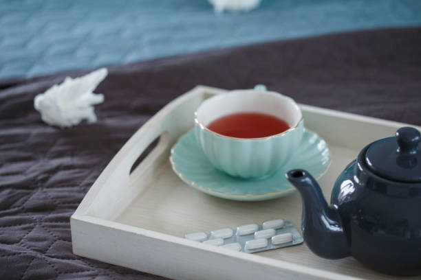 Close up shot of wooden tray lying on bed with refreshing hot cup of tea, tea cup and some prescription medication with a used tissue to fight a bad flu.