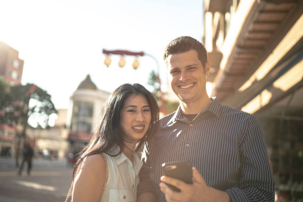 istock Happy diverse couple looking at camera and using phone in the city 1159027136