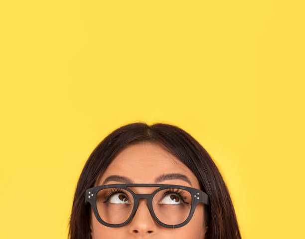 closeup portrait headshot cute happy woman in glasses looking up closeup portrait headshot cropped face above lips of cute happy woman in glasses looking up isolated on yellow studio wall background with copy space above head. Human face expressions, emotions nerd teenager stock pictures, royalty-free photos & images