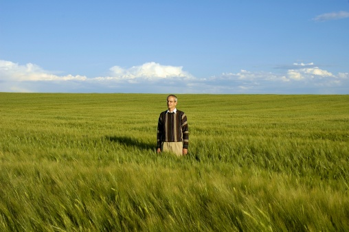A man dressed in business casual standing in a green field with blue sky and clouds on the horizon.