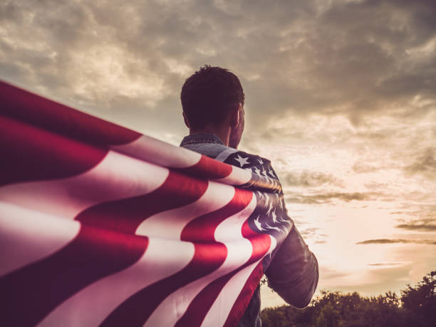 Handsome, young man waving an American flag Attractive man in jeans and a denim shirt waving an American flag against the backdrop of a sunset and a gray, gloomy sky. View from the back, close-up. National holiday concept patriotic man stock pictures, royalty-free photos & images