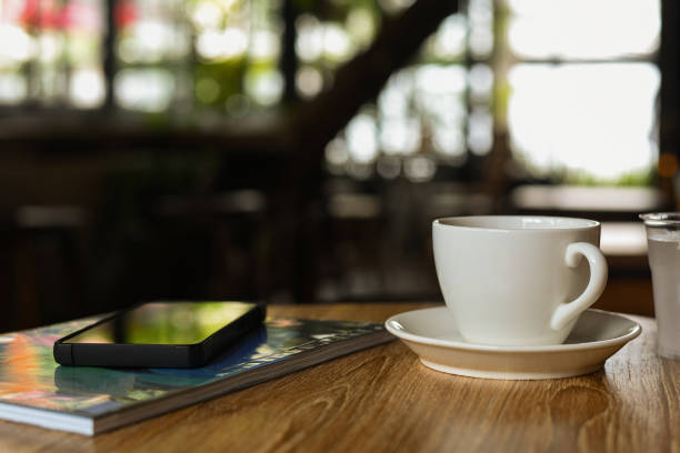 White coffee mug on wooden table with mobile phone and magazine.Blurred coffee shop background.
