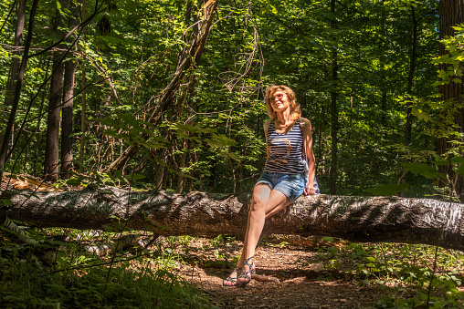 Portrait of an attractive adult woman sitting on a fallen tree on a sunny day against the background of a green forest