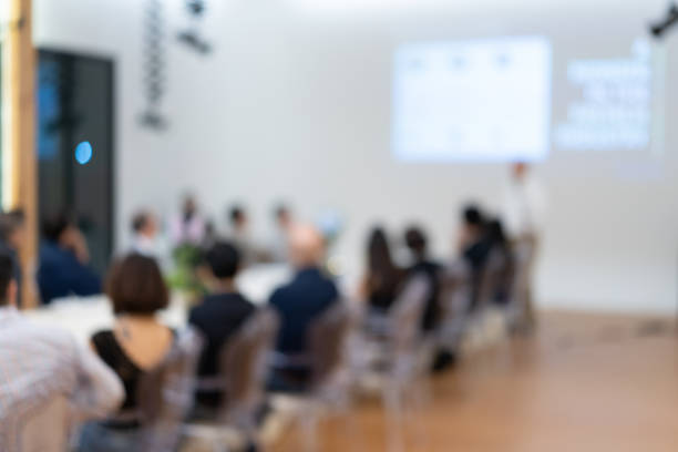 istock Blurry business presentation with the projector and white screen to the Asian audiences sit in the long table line. 1159043822