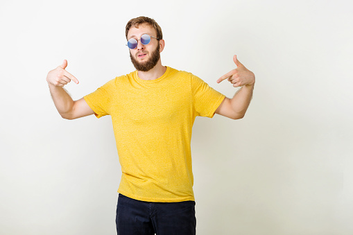You found right guy, pick me. Confident, charismatic bearded male in sunglasses promoting himself pointing at body over gray wall. Man in blank yellow t-shirt pointing at himself.
