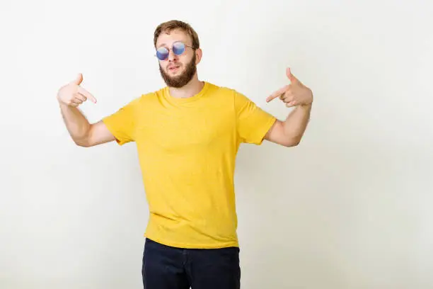 Photo of You found right guy, pick me. Confident, charismatic bearded male in sunglasses promoting himself pointing at body over gray wall. Man in blank yellow t-shirt pointing at himself.