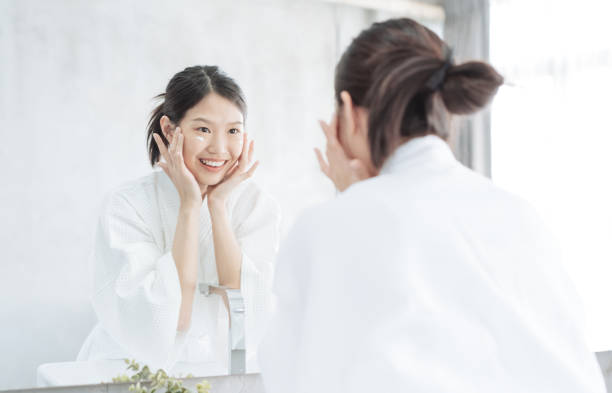 weibliche hautpflege. junge asiatische frau berührt ihr gesicht und suchen, um im badezimmer zu spiegeln - bathtub asian ethnicity women female stock-fotos und bilder