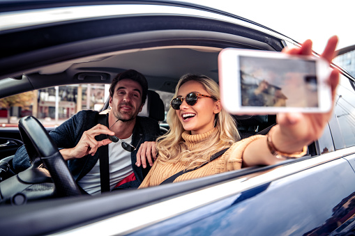 Happy young woman ready to drive car looking to her friend while she taking photo with a smartphone. Female friendship and leisure time concept.