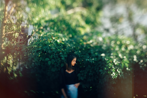 Stylish hipster girl posing at green leaves bush, atmospheric moment. Fashionable woman in denim jeans and black shirt relaxing in park. Selective focus, creative image. Space for text