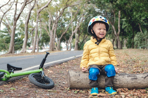 bambino asiatico di 2 - 3 anni che indossa il casco di sicurezza, seduto su un tronco di legno che si prende una pausa dopo aver guidato la sua balance bike (corri in bicicletta) nel parco - baby first steps autumn child foto e immagini stock