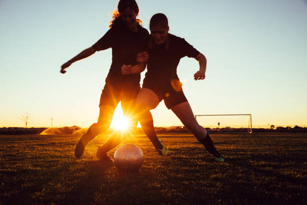 Female Soccer Players Battle for Ball Two female high school soccer players in practice uniform battle for a loose ball while on the pitch in Arizona, USA. The two teammates work on their skills with drills at dusk with the sun shining through their silhouettes. high school soccer stock pictures, royalty-free photos & images