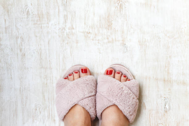 female legs with red nails in home fur fluffy pink slippers on a light wooden background. flat lay. Top view. The concept of a cozy bright girl house stock photo