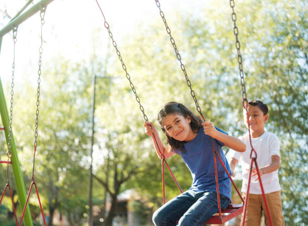 A happy girl sitting on a swing while her brother pushers her from behind.
