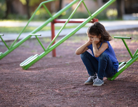 A sad little girl sitting alone on a teeter tooter with her hands on her face.