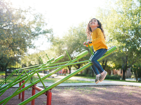 A little girl on top of a seesaw looking up and smiling on a sunny dat at the park playground.