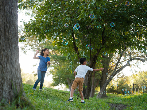 A horizontal photo of a girl and a boy standing outdoors playing with big bubbles.