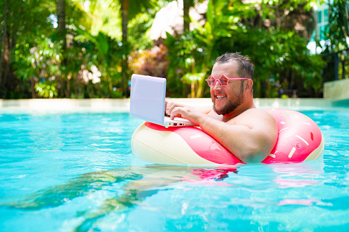 Hombre gordo divertido en vasos rosas en un círculo inflable en la piscina funciona en un ordenador portátil que retrata a una chica. photo