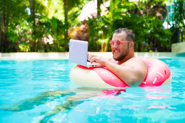 Funny fat male in pink glasses on an inflatable circle in the pool works on a laptop portraying a girl. Funny fat male in pink glasses on an inflatable circle in the pool works on a laptop portraying a girl standing water stock pictures, royalty-free photos & images