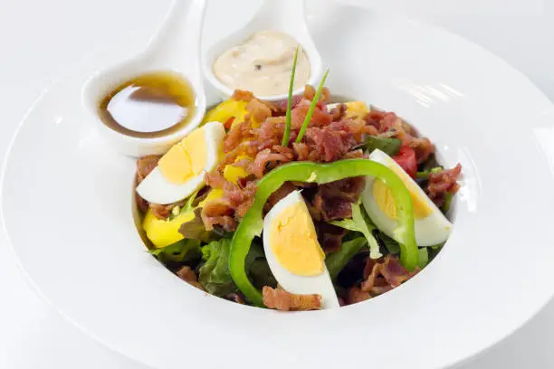 Fresh Caesar Salad with Deep-fried Bacon and Boiled Eggs & Salad-dressing on White Porcelain Round Plate, on White Background with Shadow. Close-Up High Angle Side View, Selective Focus at Food.