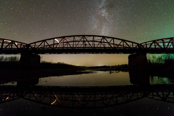 istock Illuminated metal bridge on concrete supports reflected in water on dark starry sky with Milky Way constellation background. Night photography concept. 1159167360