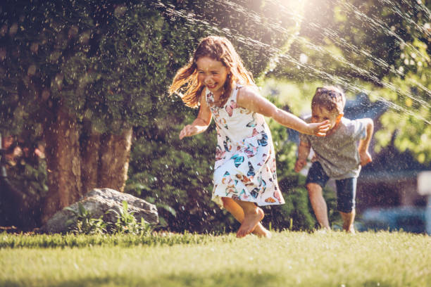 niños felices jugando con el aspersor de jardín - saltar actividad física fotos fotografías e imágenes de stock