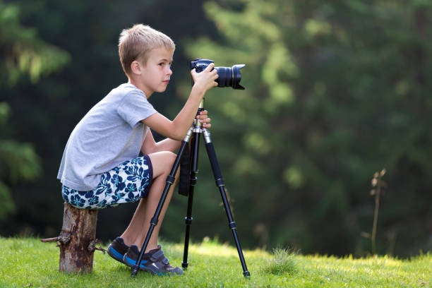 istock Young blond child boy sitting on tree stump on grassy clearing taking picture with tripod camera. 1159180749