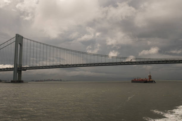 On a stormy morning a tugboat is pushing a barge under the Verrazano Narrows Bridge