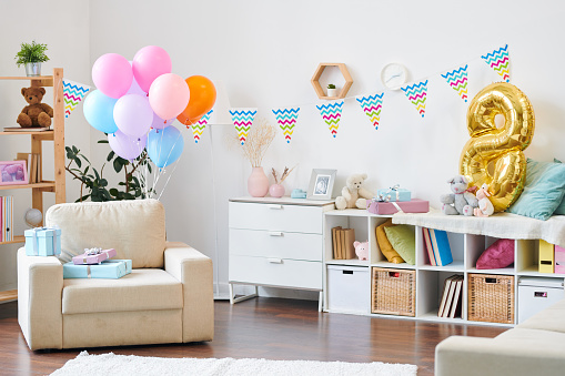 Interior of living-room in modern flat prepared for birthday party of little kids and decorated with balloons and flags