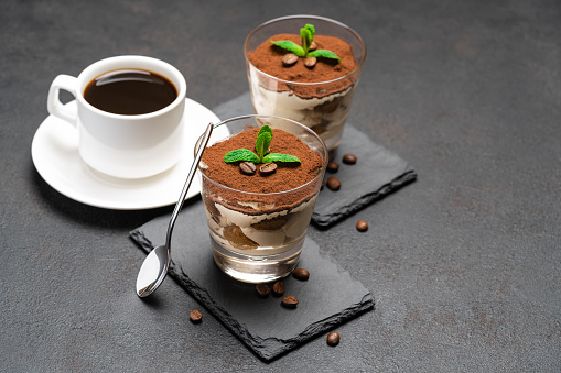 Classic tiramisu dessert in a glass on stone serving board and cup of coffee on dark concrete background or table