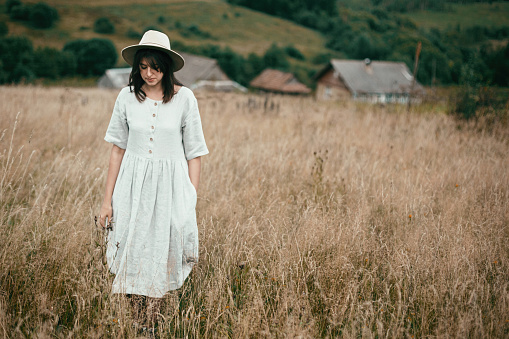 Stylish girl in linen dress and hat walking among herbs and wildflowers in field. Boho woman enjoying day in countryside, simple slow life style. Space for text. Atmospheric image