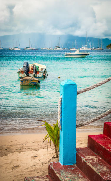 Red painted old stone staircase with cyan blue pole and thick cord on the beach. Fishing boat on the water. In the background rain clouds. Terre-de-Haut, Guadeloupe.