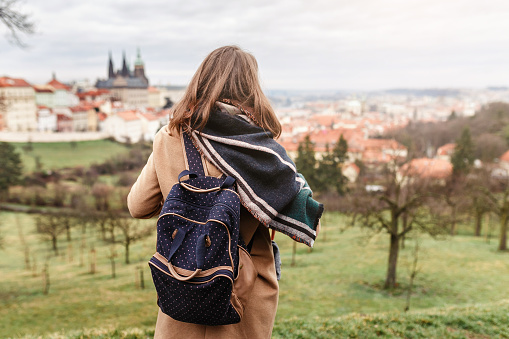 Woman tourist in a coat with a backpack travels in the city park of Prague, rear view