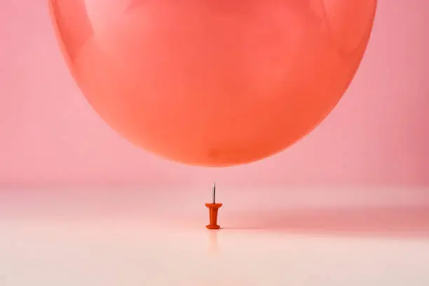 Photo of Red balloon fall on a pin needle on pink background. Danger or protection concept