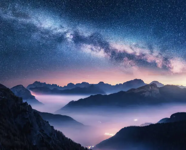 Milky Way over mountains in fog at night in summer. Landscape with foggy alpine mountain valley, purple low clouds, colorful starry sky with milky way, city illumination. Dolomites, Italy. Space