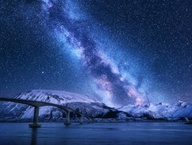 istock Bridge and starry sky with Milky Way over snow covered mountains reflected in water. Night landscape with road, snowy rocks, purple sky with stars and milky way, sea. Winter in Lofoten islands, Norway 1159272367