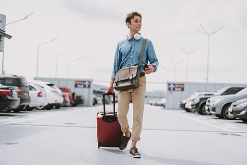 Full length portrait of young guy in headphones holding suitcase and smartphone while going along the parking outdoors. Copy space in left side