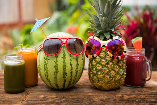 This is a photograph of a watermelon and pineapple with sunglasses sitting on a wooden picnic bench symbolizing summertime fun, tropical food and healthy eating