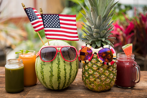 This is a photograph of a watermelon and pineapple with sunglasses sitting on a wooden picnic bench symbolizing summertime fun, tropical food and healthy eating