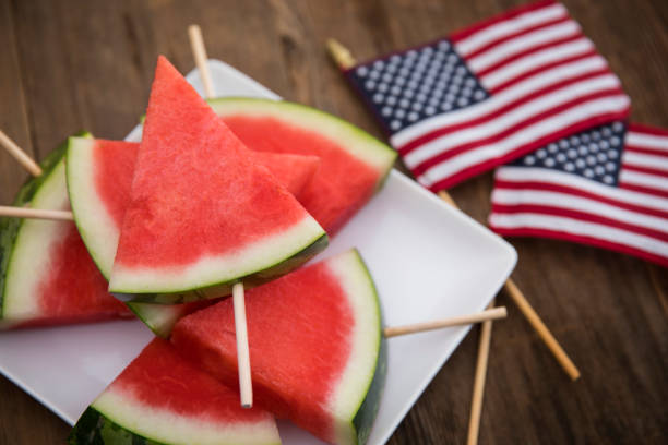 This is a high angle photograph of a plate of triangle shaped sliced cut watermelon with popsicle sticks making it easier to eat on a white modern plate. It is sitting on a wood Beach deck