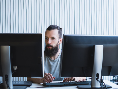 Quality assurance. Bearded software engineer using two monitors, working on task at office. Copy space.