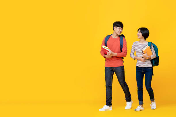 Students holding book and talking while walking Asian girl and boy students with bags holding books in a conversation while walking towards class college isolated against yellow studio background male chinese student stock pictures, royalty-free photos & images
